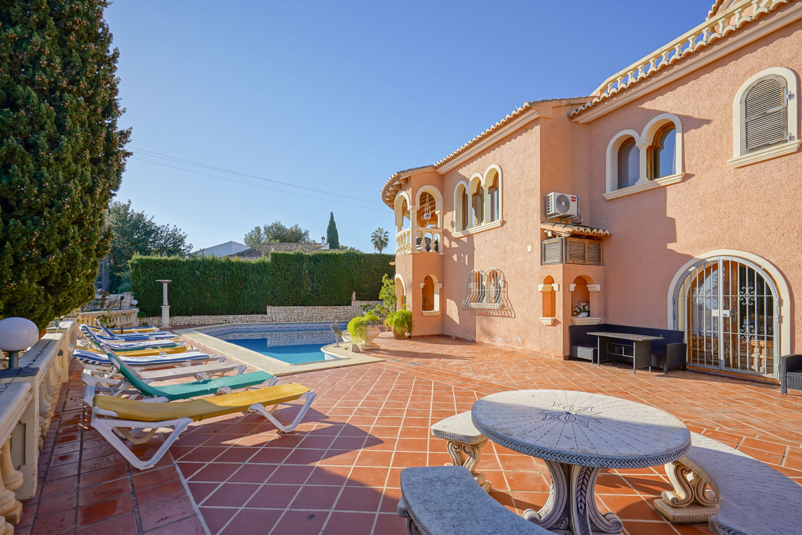 Amplia terraza de baldosas de terracota con piscina, zona de tumbonas y mesa de piedra tallada bajo una fachada de estilo mediterráneo con arcos.