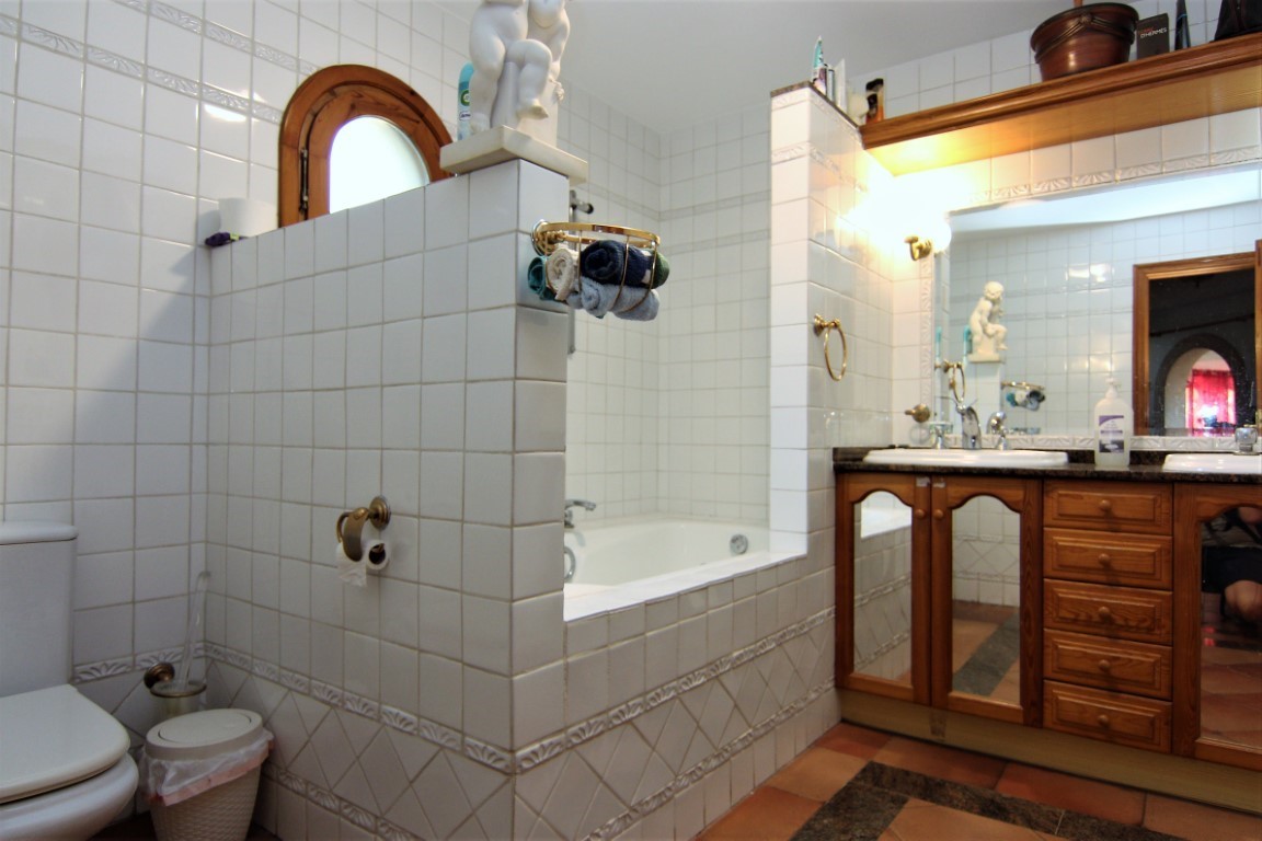 Baño con paredes alicatadas en blanco, bañera encastrada y mueble de lavabo doble en madera con encimera de granito. Destaca el suelo de terracota y la ventana de arco.