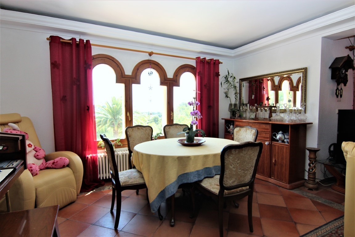 Comedor con mesa redonda, sillas tapizadas y un ventanal de madera con tres arcos. Cuenta con suelo de baldosas de terracota y un aparador clásico con espejo.