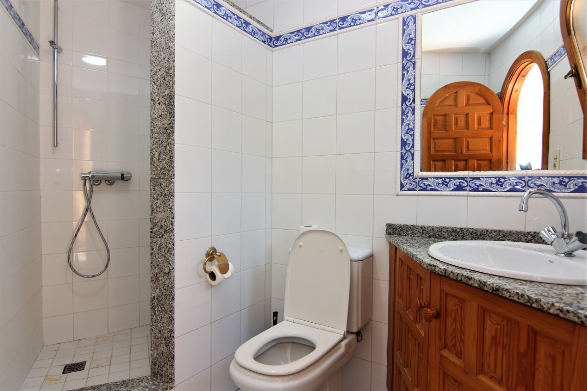 Cuarto de baño con ducha de obra, encimera de granito, mueble de madera y azulejos blancos con cenefa decorativa azul.