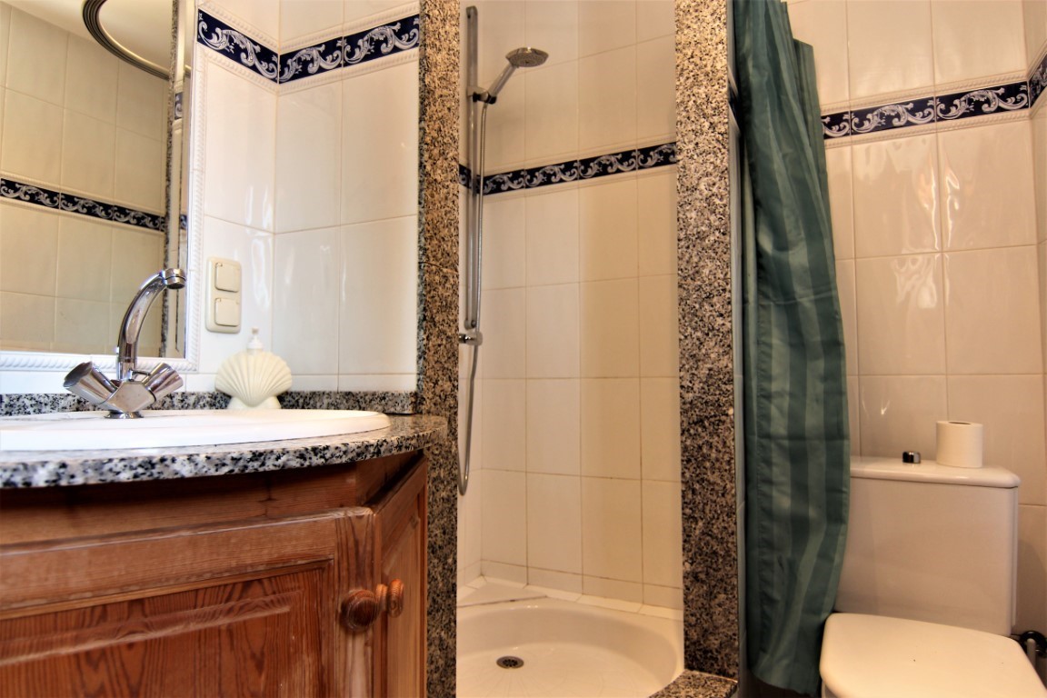 Cuarto de baño con encimera de granito, mueble de madera, ducha con marco de piedra y azulejos blancos con cenefa decorativa azul.