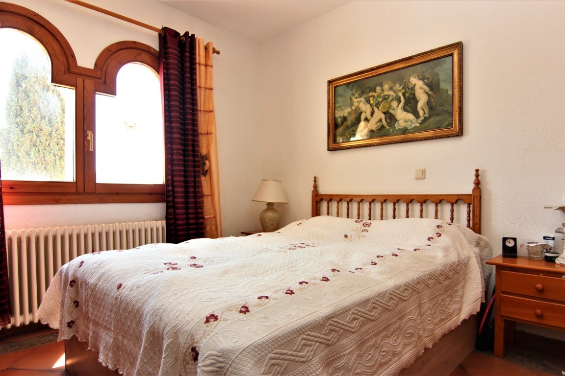 Dormitorio con cama doble, cabecero de madera torneada, ventana de arco doble con marcos de madera y un cuadro clásico sobre la pared blanca.