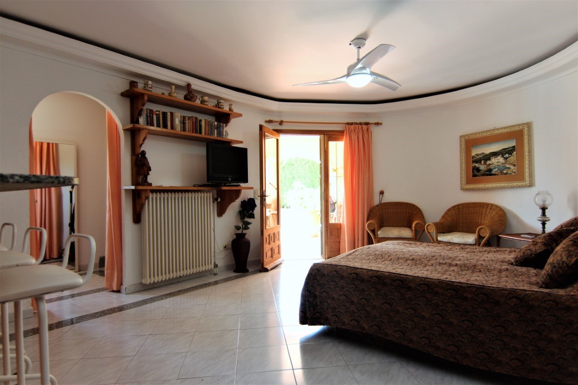 Dormitorio espacioso con acceso directo a la terraza, zona de estar con sillones de mimbre, estanterías de madera y ventilador de techo sobre suelo cerámico.