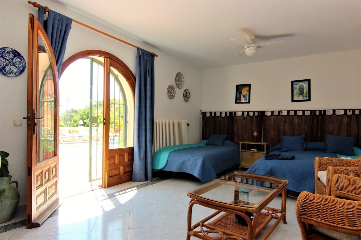 Dormitorio luminoso con acceso directo a la terraza mediante un gran ventanal de arco en madera, mobiliario de mimbre y suelos cerámicos claros.
