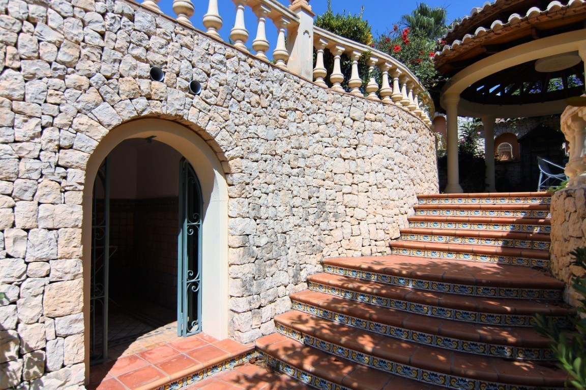 Escalera exterior de terracota con azulejos decorativos junto a un muro curvo de piedra natural. Incluye un arco de acceso y balaustrada clásica superior.