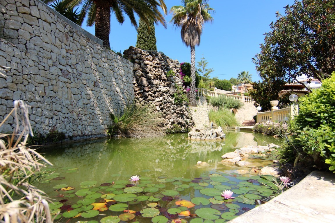 Estanque ornamental con nenúfares y cascada de roca natural, rodeado de muros de piedra seca y vegetación mediterránea con palmeras.