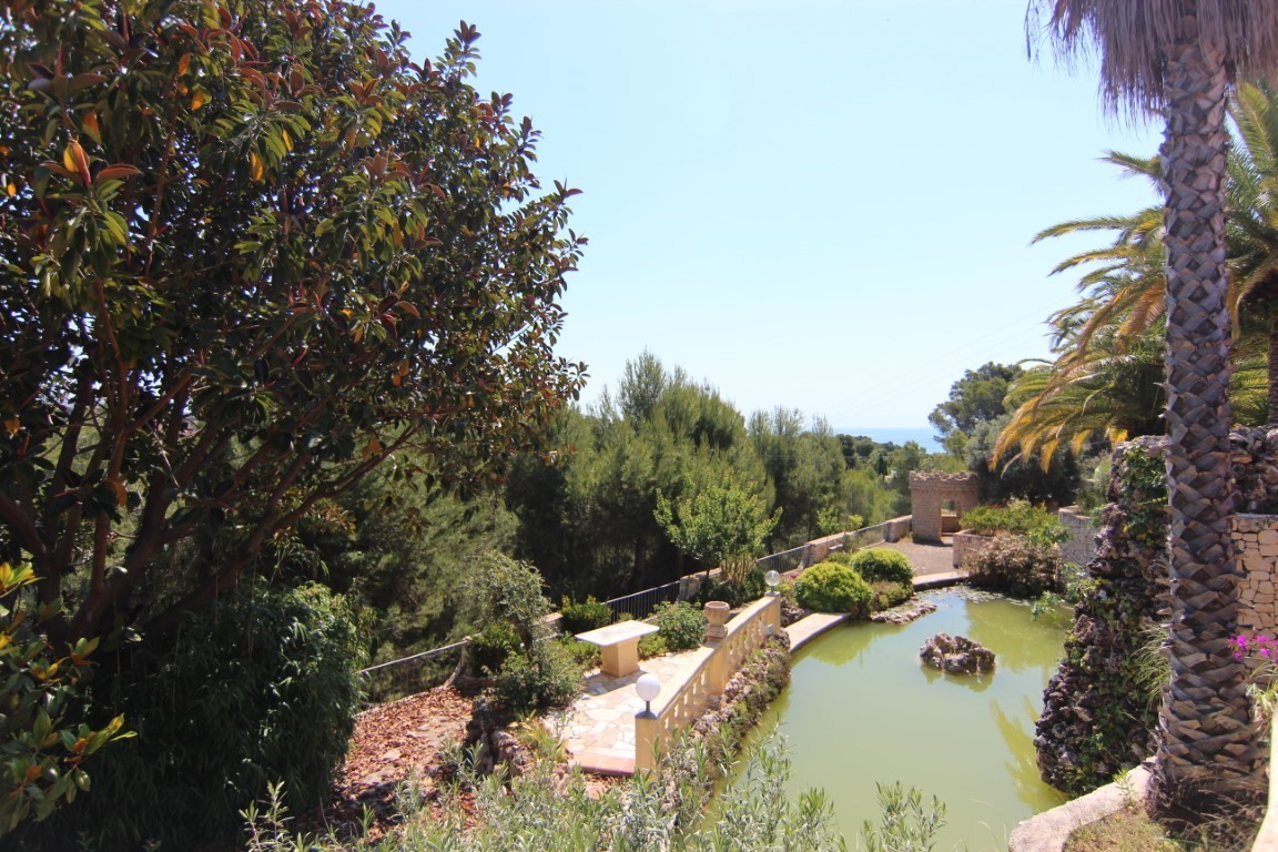 Gran estanque ornamental con cascada y elementos de piedra, rodeado de vegetación mediterránea y con vistas parciales al mar desde la parcela.