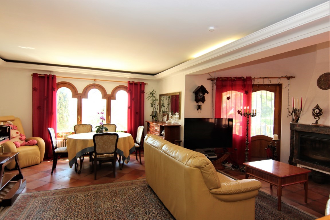 Salón-comedor con suelos de terracota, chimenea de mármol y ventanales de arco de madera. La estancia cuenta con iluminación indirecta en el techo y acceso directo al exterior.