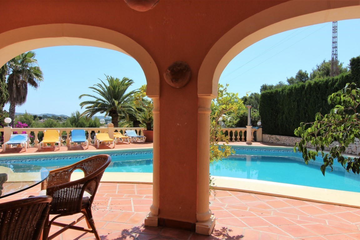Terraza cubierta con arcos mediterráneos que ofrece vistas a la piscina y al solárium equipado con tumbonas, rodeada de vegetación y palmeras.