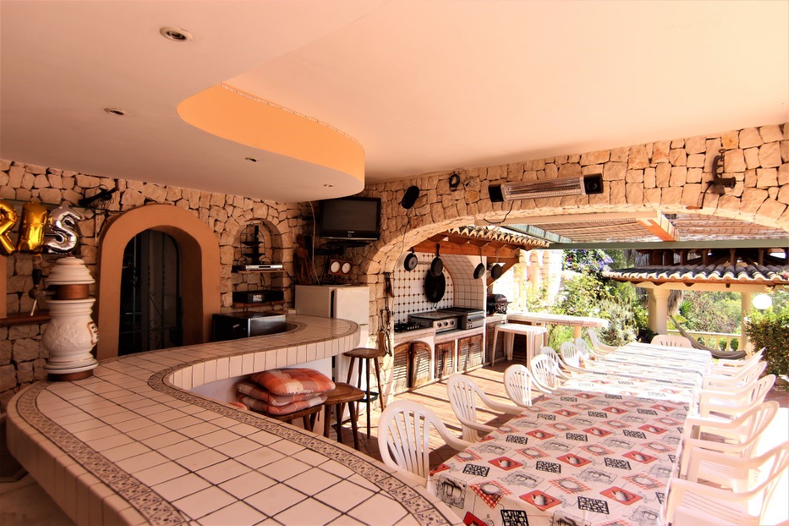Terraza cubierta con bar de azulejos, cocina exterior profesional y zona de comedor amplia bajo arcos de piedra natural.