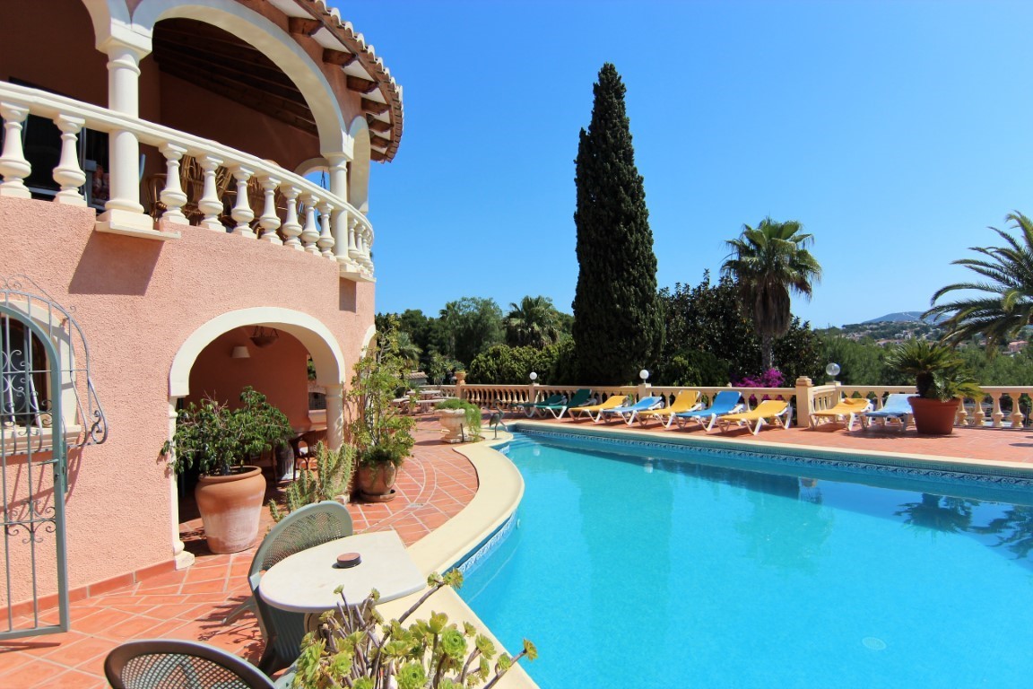 Terraza de terracota con piscina curva, tumbonas alineadas y fachada mediterránea con arcos clásicos y balaustradas blancas bajo un cielo despejado.