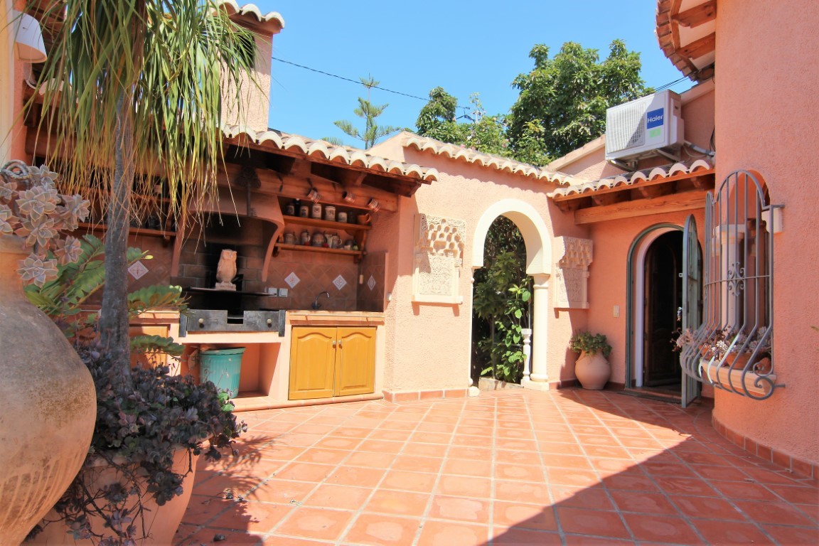 Terraza exterior con cocina de verano, barbacoa de obra y suelos de terracota. Destaca el arco de estilo mediterráneo y la carpintería de madera bajo el porche.