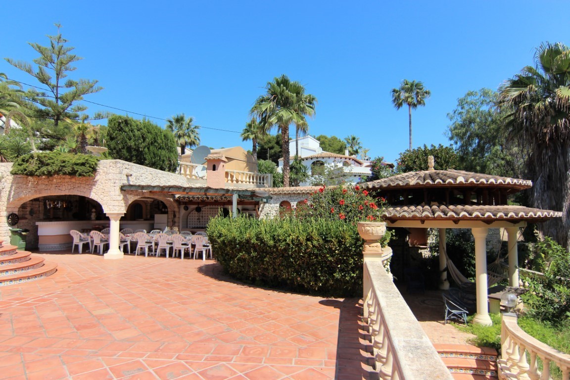 Terraza exterior con suelos de terracota, zona de comedor bajo arcos de piedra y un cenador con tejado de tejas rodeado de vegetación mediterránea.