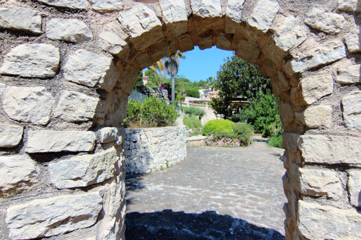 Un arco de piedra natural enmarca la entrada a los jardines mediterráneos, con senderos empedrados y muros de contención de piedra seca.