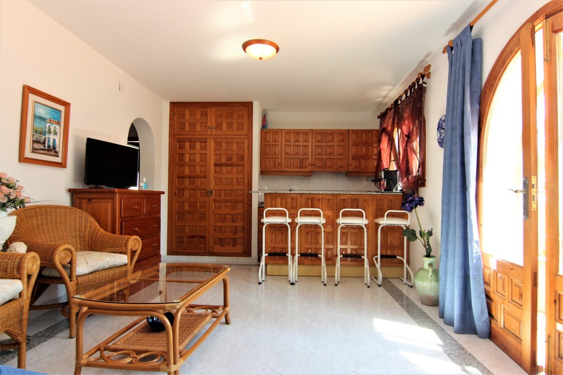 Zona de estar de concepto abierto con muebles de mimbre, cocina integrada con barra de madera y armarios empotrados con paneles decorativos.