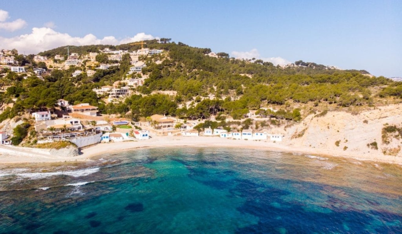 Vista aérea de la Cala de la Barraca con sus icónicas casas blancas de puertas azules, aguas cristalinas y laderas boscosas en Jávea.