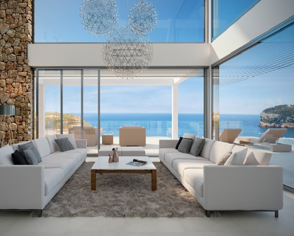 Salón de doble altura con ventanales de suelo a techo, pared de piedra natural y lámparas de diseño. Ofrece vistas panorámicas al mar y acceso a la terraza.