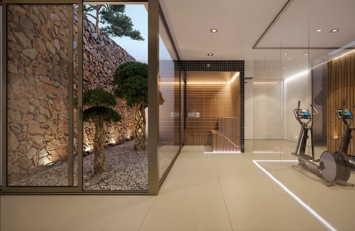 Zona de bienestar con gimnasio acristalado y sauna de madera, conectada a un patio exterior con muro de piedra natural y vegetación decorativa.