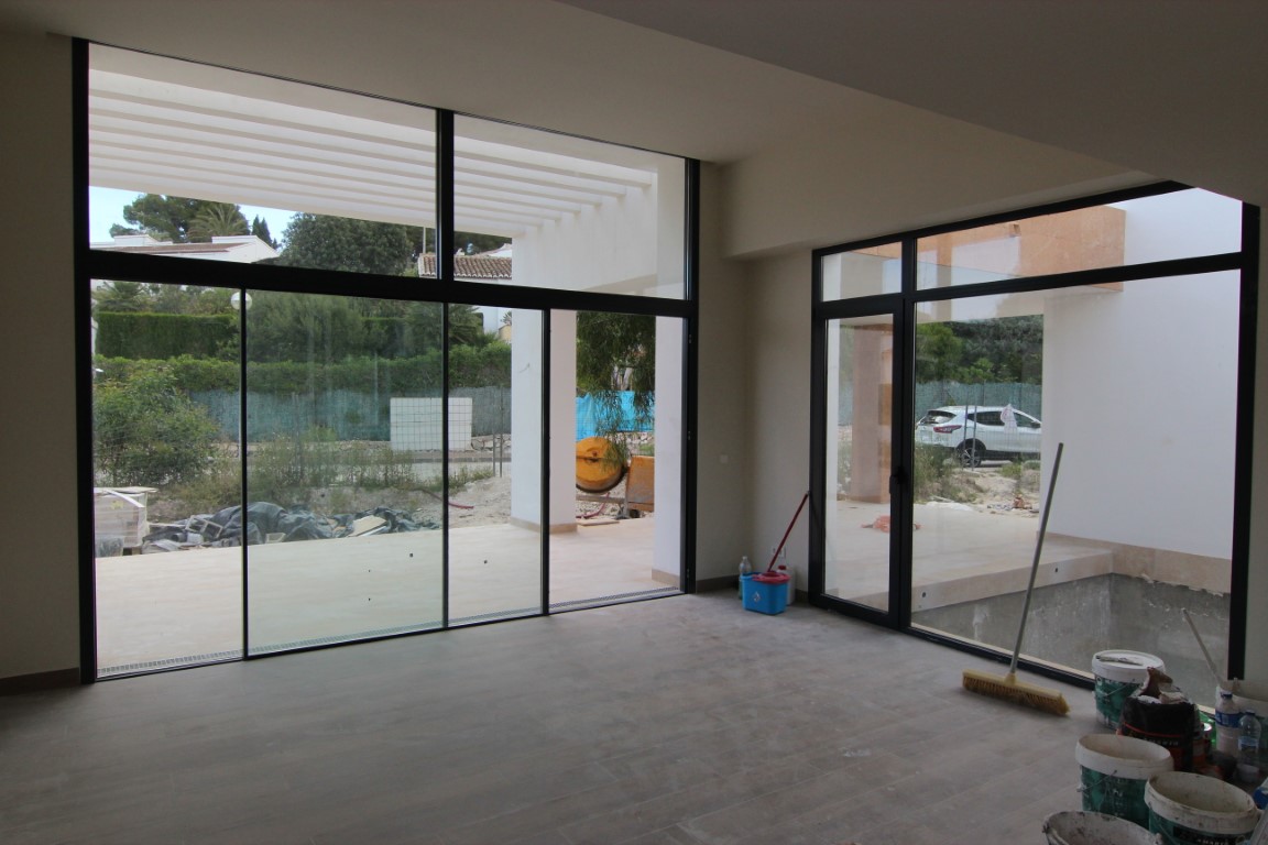 Salón en fase de construcción con amplios ventanales de carpintería negra que dan acceso a la terraza con pérgola blanca y vistas al exterior.