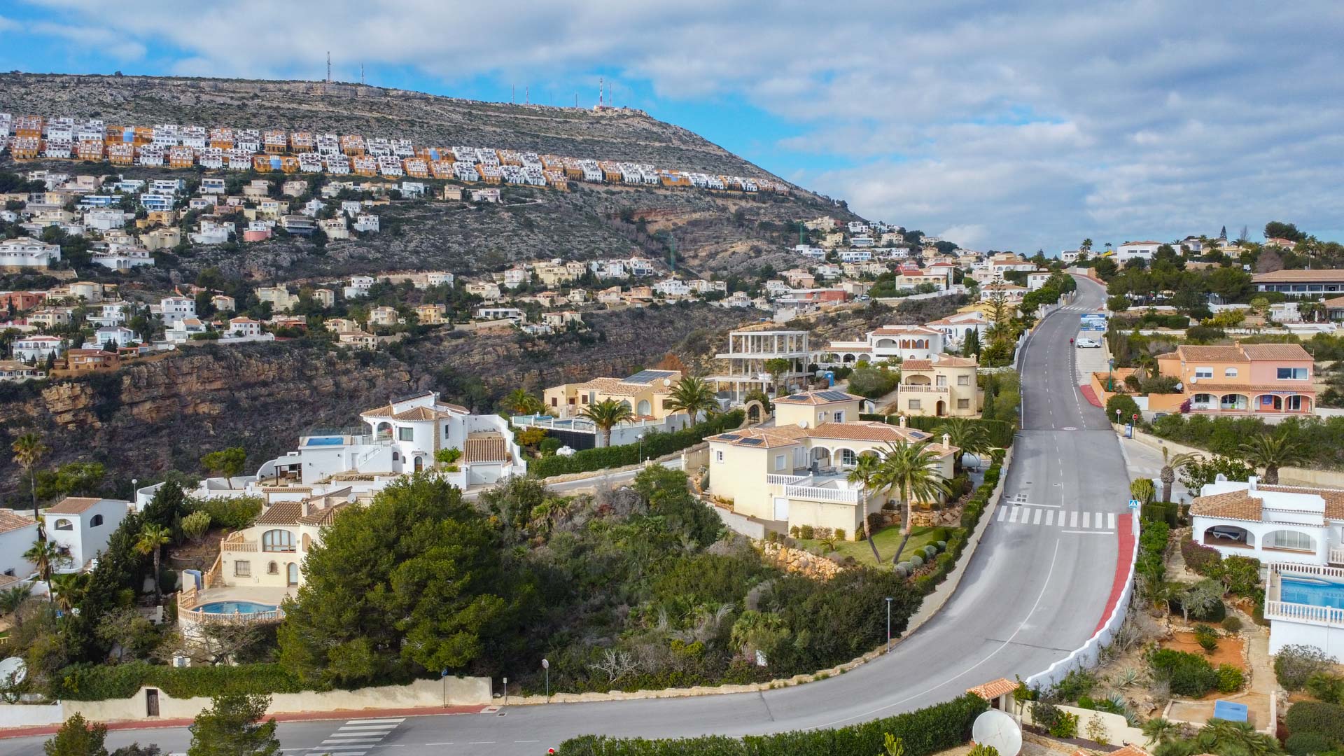 Vista aérea del entorno residencial en Cumbre del Sol, destacando la carretera principal, villas de estilo mediterráneo y la ladera urbanizada bajo un cielo despejado.