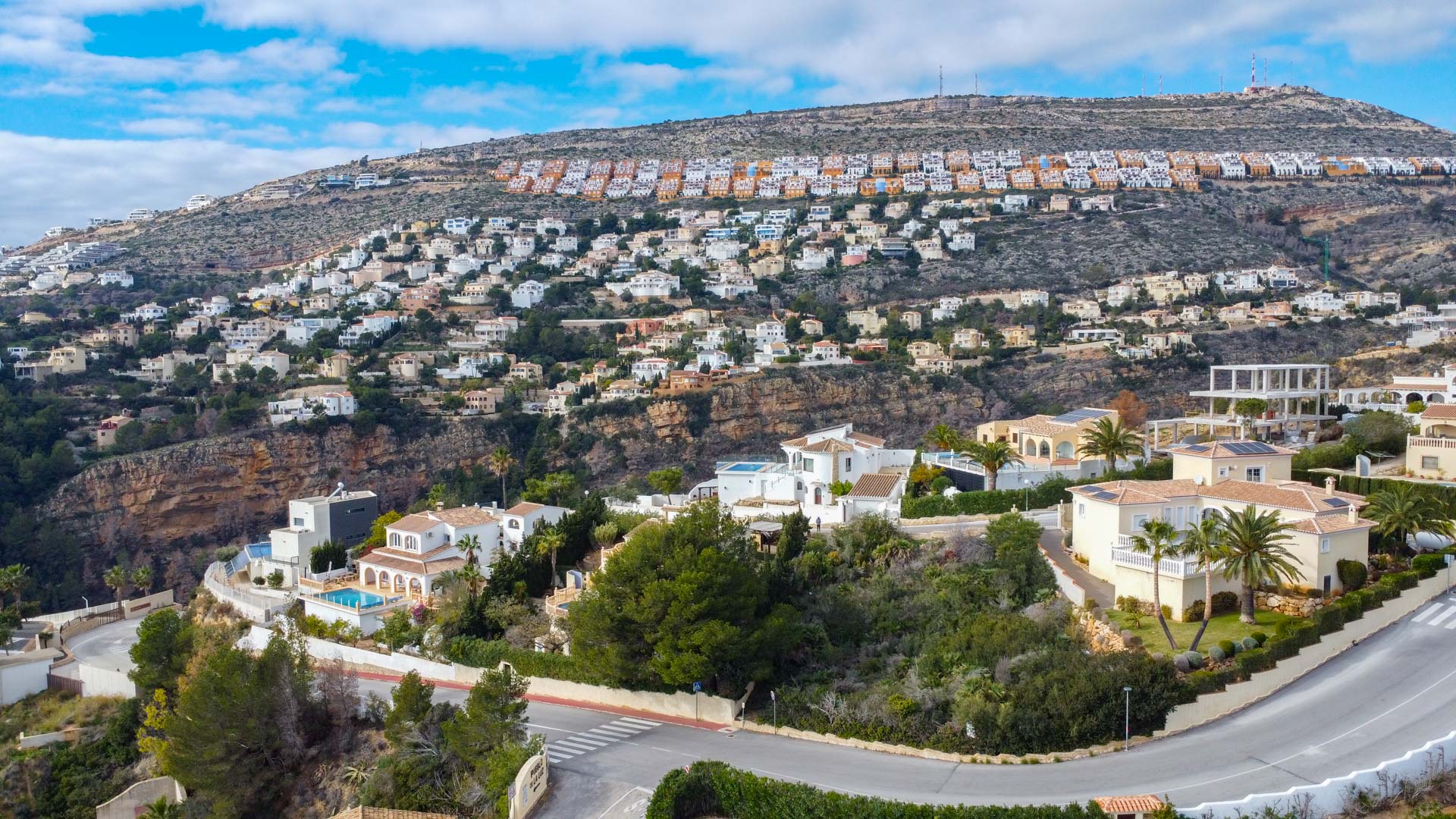 Vista aérea panorámica de la urbanización en Cumbre del Sol, mostrando villas unifamiliares integradas en la ladera con vegetación mediterránea y carreteras sinuosas.