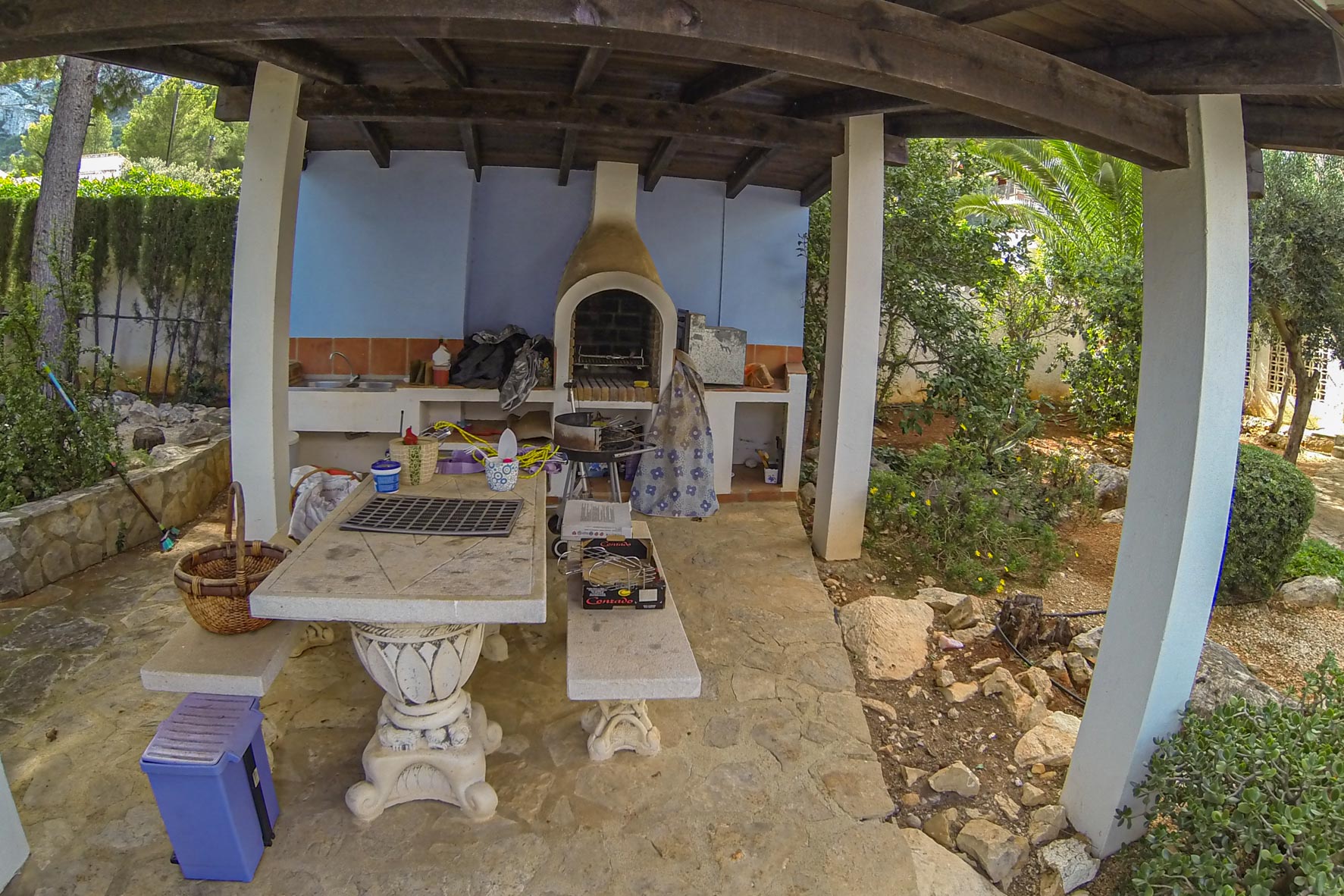Barbacoa cubierta con horno de ladrillo y mesa de piedra en jardín exuberante.