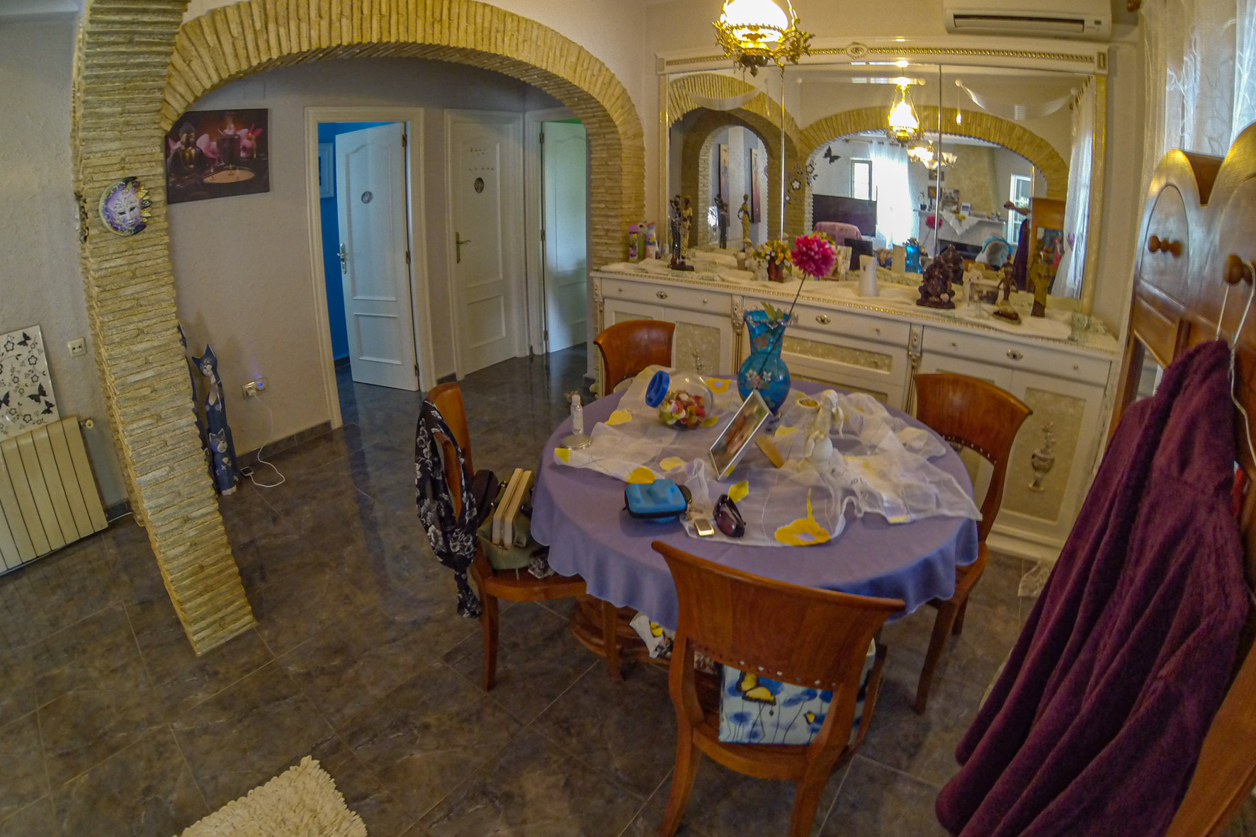 Comedor con arcos de piedra, mesa redonda con mantel lila y aparador blanco con espejo ornamentado.