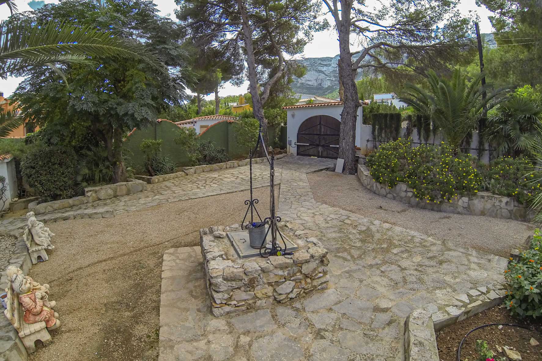 Entrada a la propiedad con pozo de piedra, camino empedrado y portón de madera al fondo.