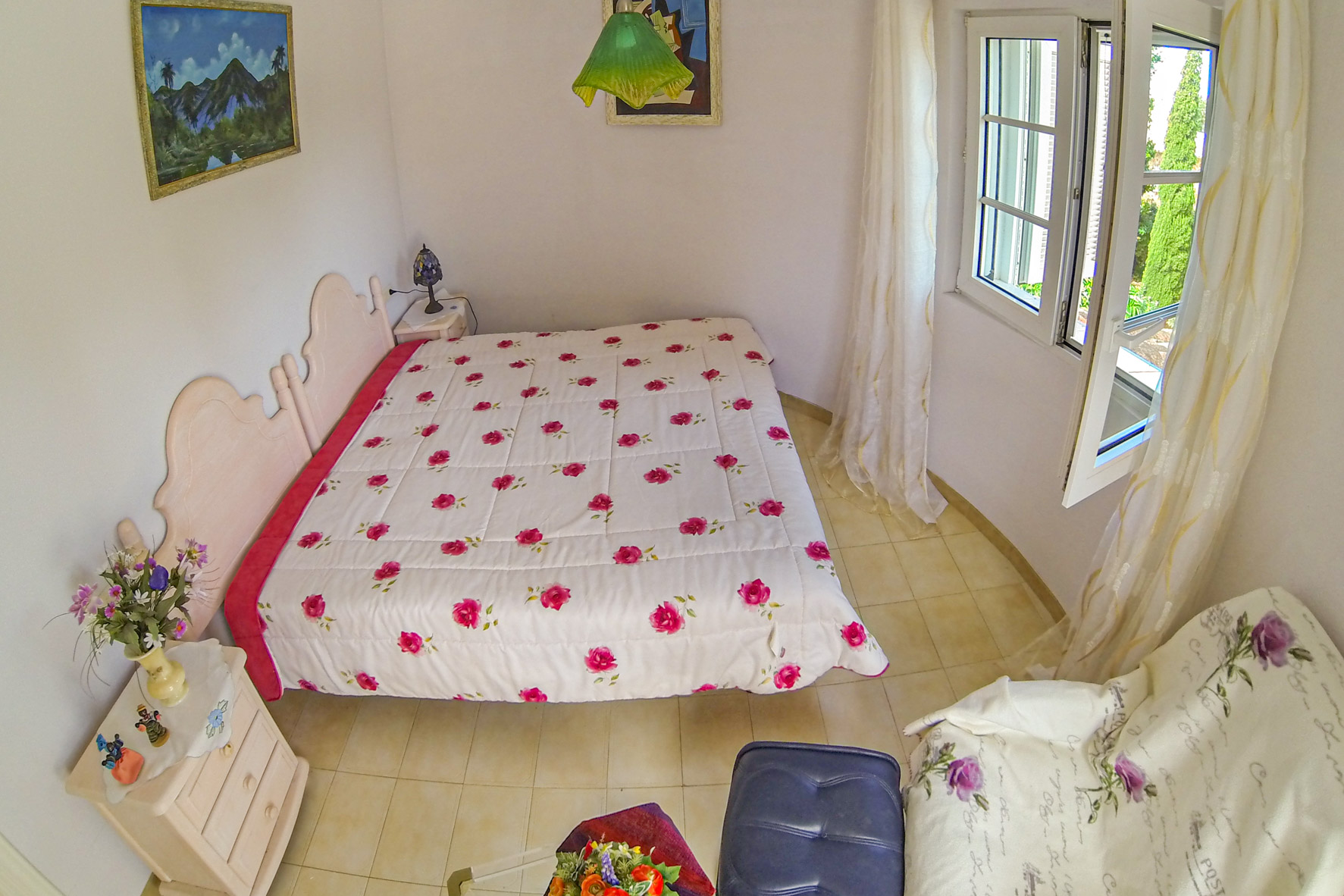 Habitación luminosa con cama doble, ropa de cama floral y ventana con cortinas traslúcidas.