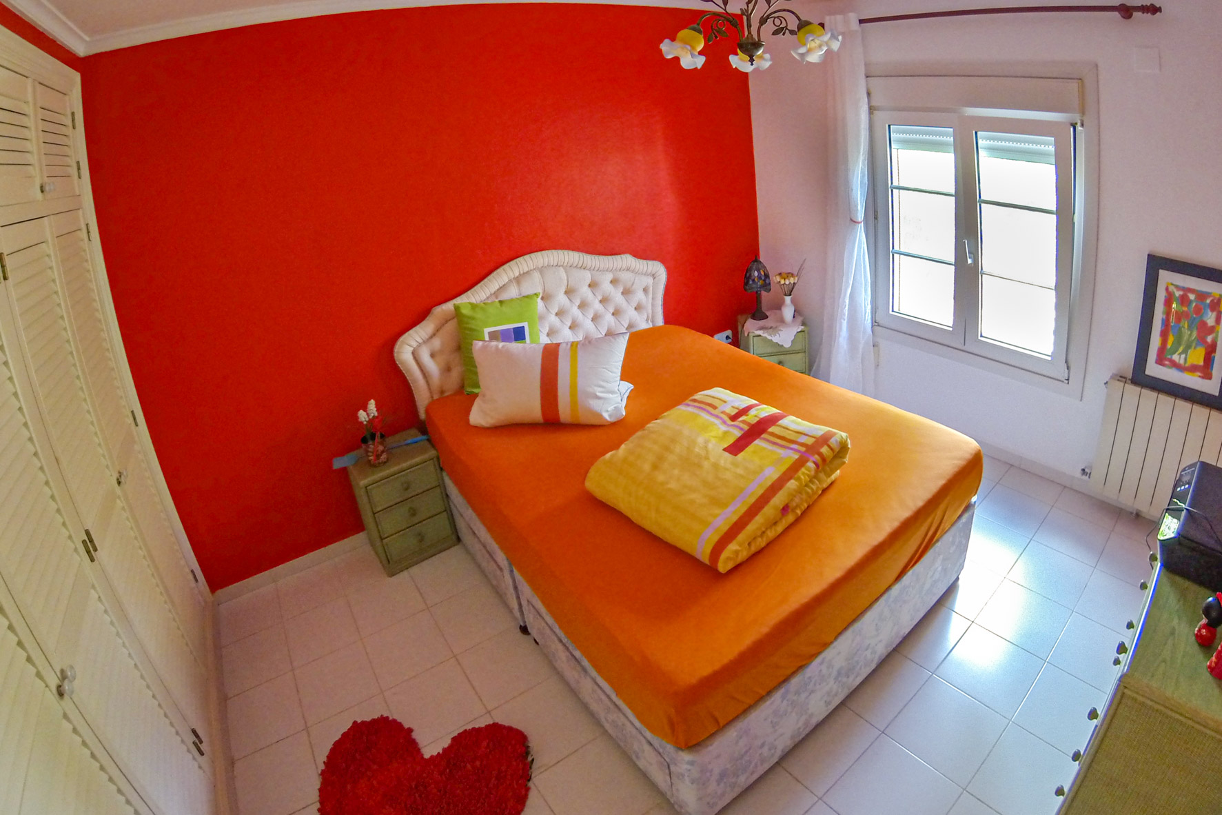 Habitación luminosa con pared roja, cama doble con ropa de cama naranja y cabecero tapizado.