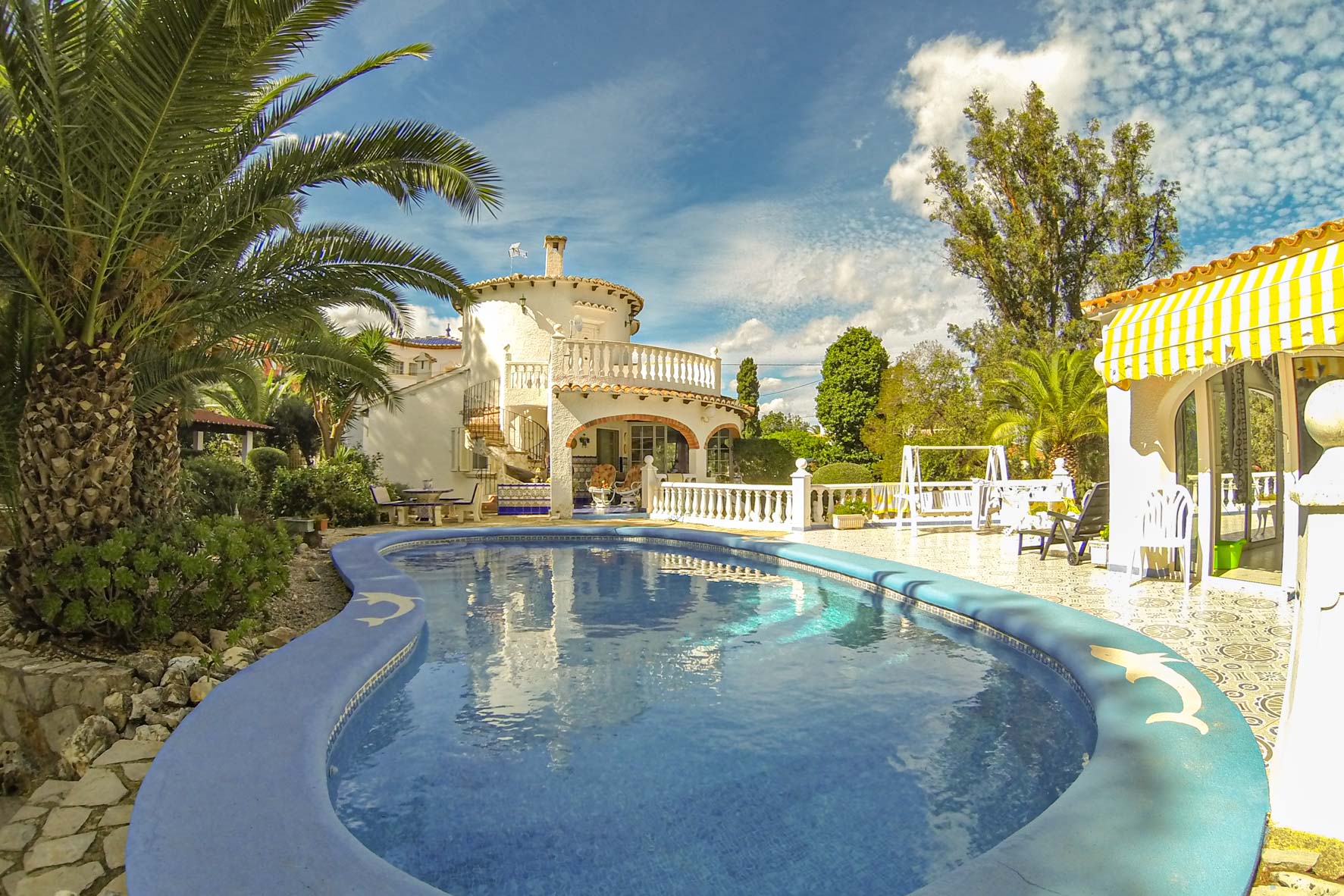 Vista de la piscina azul turquesa con borde decorado con delfines y la fachada de la villa blanca de dos pisos.