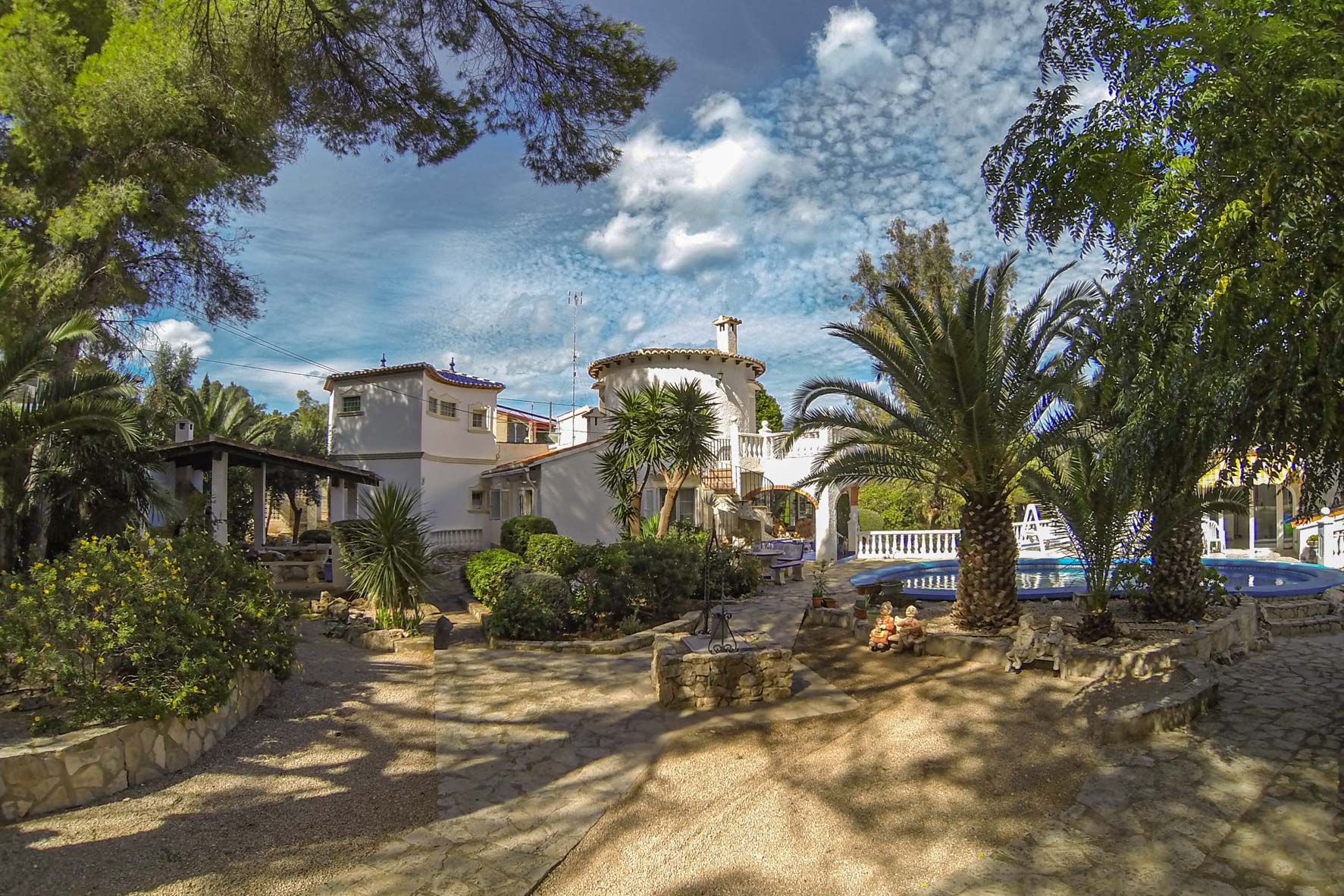 Vista del jardín maduro con piscina circular y casa principal de estilo mediterráneo.