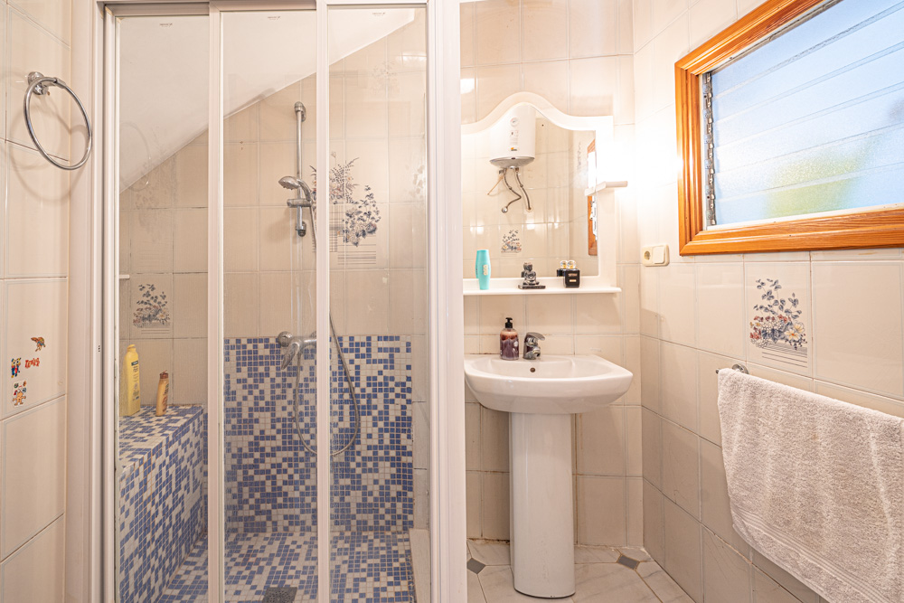 Cuarto de baño con ducha de obra revestida en mosaico azul, lavabo de pedestal y paredes alicatadas con detalles florales y ventana de madera.