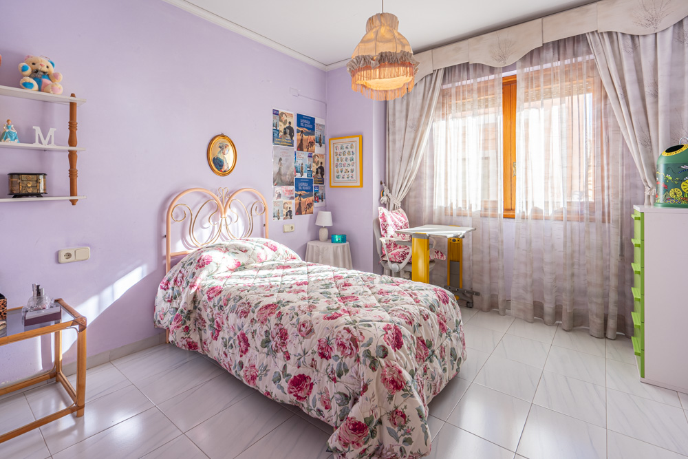 Dormitorio individual con paredes lavanda, suelo cerámico claro y cama con cabecero de forja. Incluye un escritorio junto a la ventana de madera que aporta luz natural.