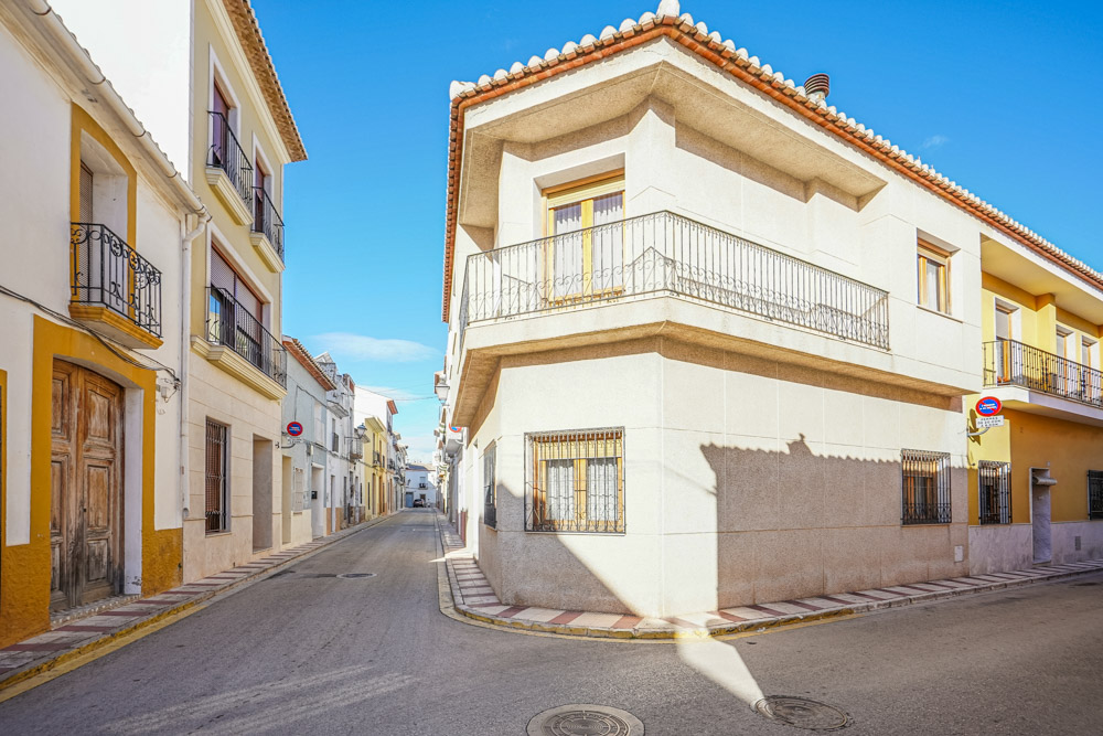 Exterior de la casa de pueblo situada en una esquina, destacando su fachada clara, balcones con herrería y ubicación en una calle tranquila del centro de Gata de Gorgos.