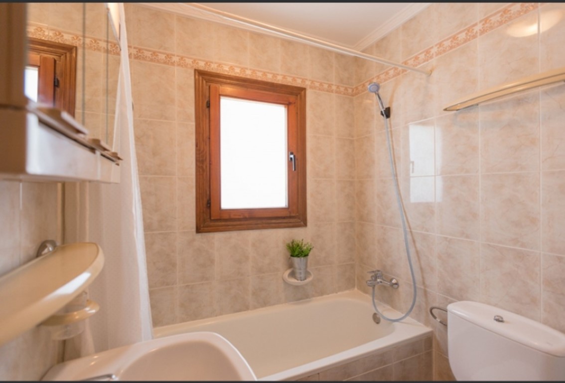 Cuarto de baño funcional con bañera, paredes alicatadas en tonos beige con cenefa decorativa y una ventana de madera que aporta luz natural.