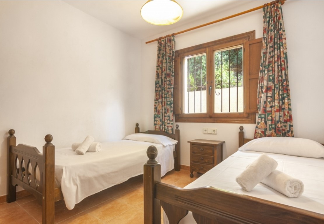 Dormitorio con dos camas individuales de madera, suelos de terracota y ventana con marco de madera que ofrece luz natural y vistas al exterior.