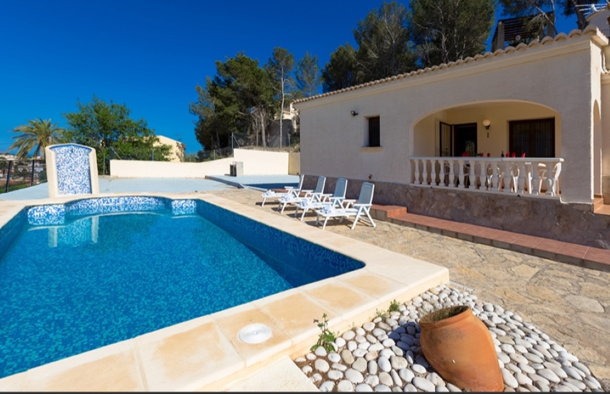 Exterior de la villa con piscina de mosaico azul, solárium de piedra natural y terraza cubierta con balaustrada blanca bajo un cielo despejado.
