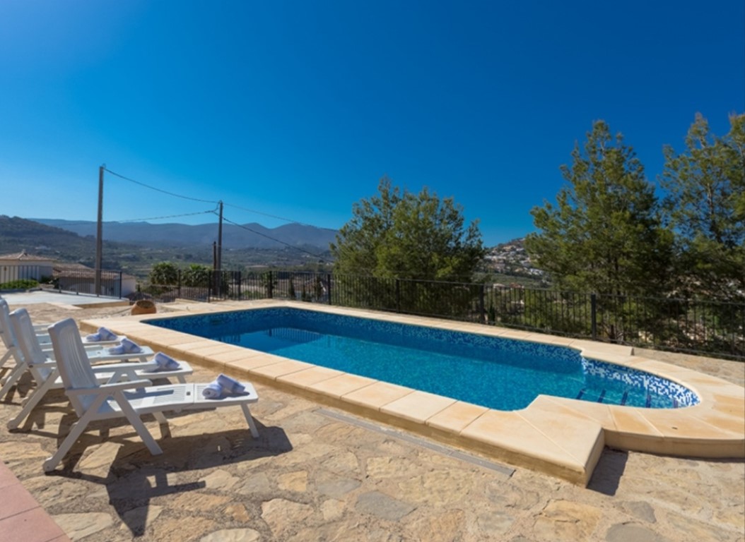 Piscina rectangular con mosaico azul y escalera lateral, rodeada por una terraza de piedra natural con tumbonas y vistas despejadas a las montañas.