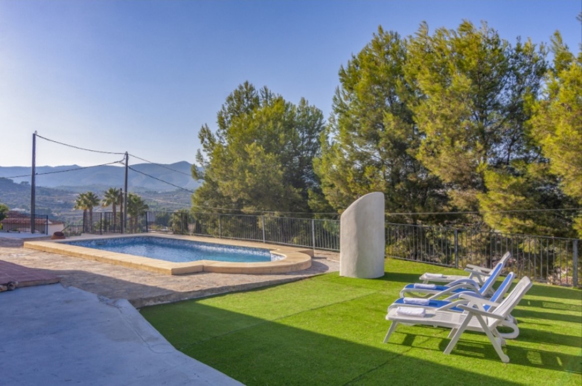 Zona de piscina exterior con terraza de piedra, césped artificial y tumbonas, rodeada de pinos maduros y vistas despejadas a las montañas.