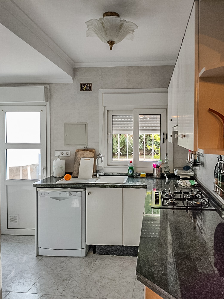 Cocina funcional con encimera de granito oscuro, equipada con placa de gas, lavavajillas y acceso directo al exterior. Incluye mobiliario blanco y suelos cerámicos.