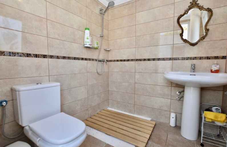 Cuarto de baño con ducha a ras de suelo, plato de madera, azulejos beige con cenefa decorativa y espejo de estilo clásico con marco dorado.