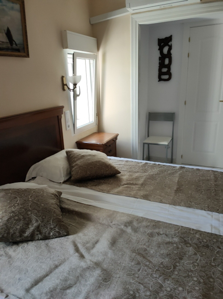Dormitorio con cama doble, cabecero de madera clásica y aire acondicionado. Dispone de ventana con luz natural, mesita de noche y un rincón con silla auxiliar junto a la puerta.