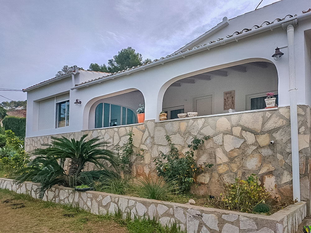 Exterior de la villa con una amplia naya arqueada y fachada de piedra natural. El jardín delantero presenta vegetación mediterránea sobre un muro de mampostería.