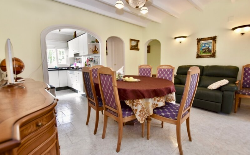 Salón-comedor de la casa de invitados con vigas vistas, mobiliario clásico y un arco que conecta con la cocina blanca equipada.