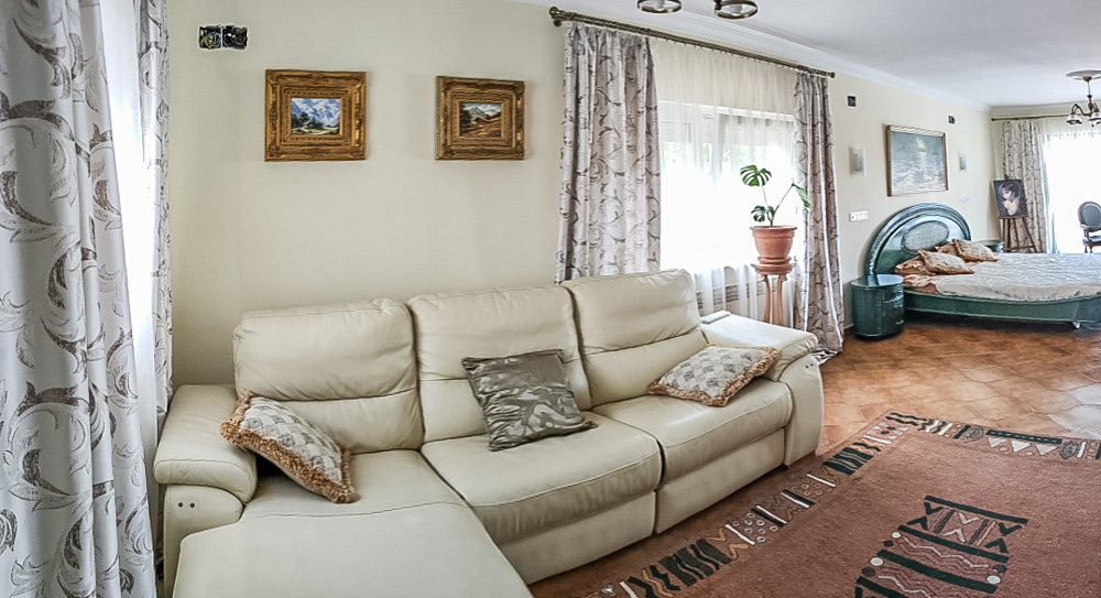 Salón de la casa de invitados con sofá de piel color crema, suelos de baldosa cerámica y zona de dormitorio integrada con mobiliario de estilo clásico.