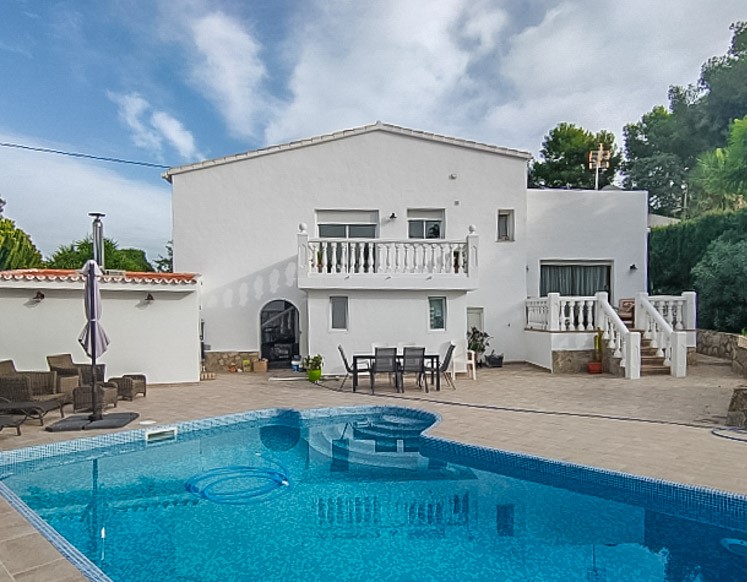 Vista exterior de la piscina de mosaico azul integrada en una amplia terraza de gres, con la fachada blanca de la villa y zonas de comedor al aire libre.