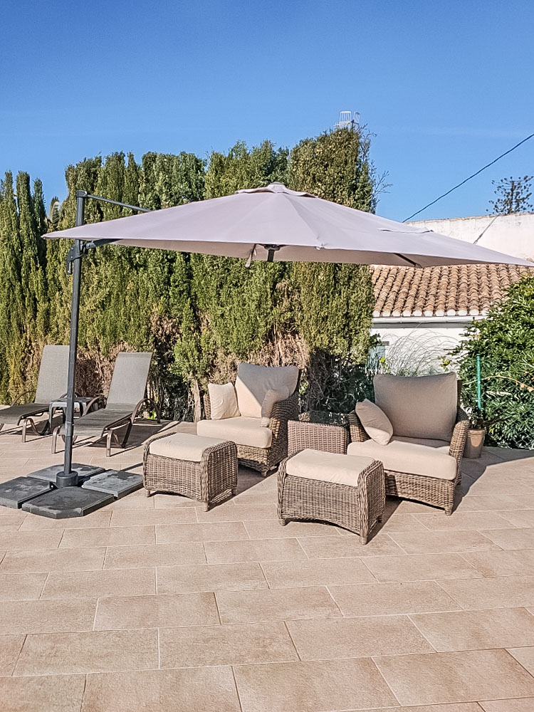 Zona de descanso exterior con sillones de mimbre sintético, reposapiés y sombrilla lateral sobre suelo de gres cerámico, rodeada de setos altos.