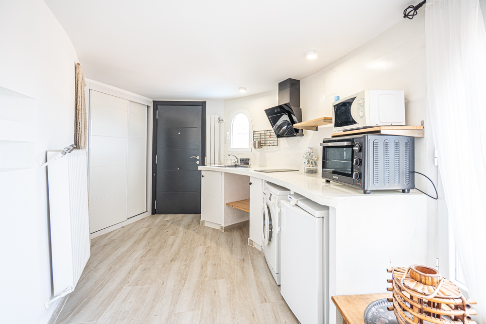 Cocina compacta renovada con encimera blanca curva, equipada con electrodomésticos modernos, suelos de gres efecto madera y acceso independiente.