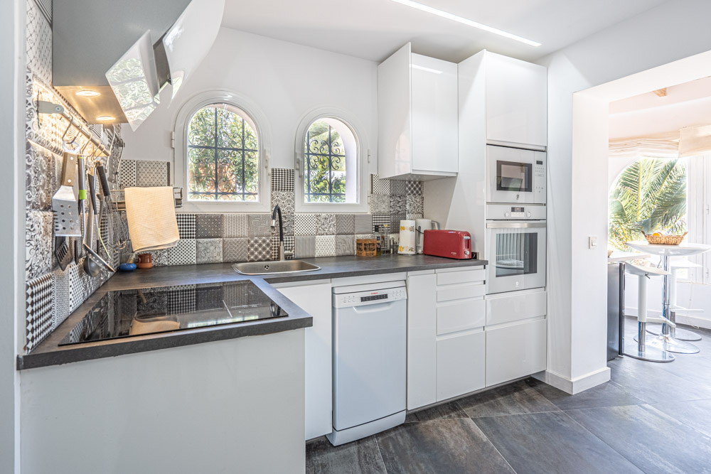 Cocina moderna con armarios blancos brillantes, salpicadero de azulejos decorativos y electrodomésticos integrados. Ventanas de arco proporcionan luz natural y vistas al jardín.